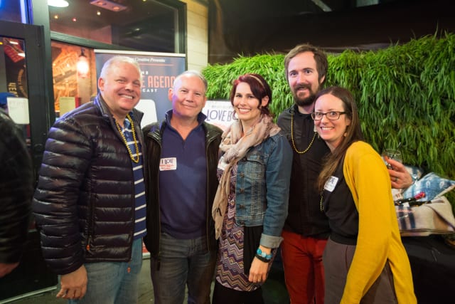Group of attendees at Love Bomb 2017 event, posing together with smiles, wearing festive attire and beads, with greenery and event signage in the background.